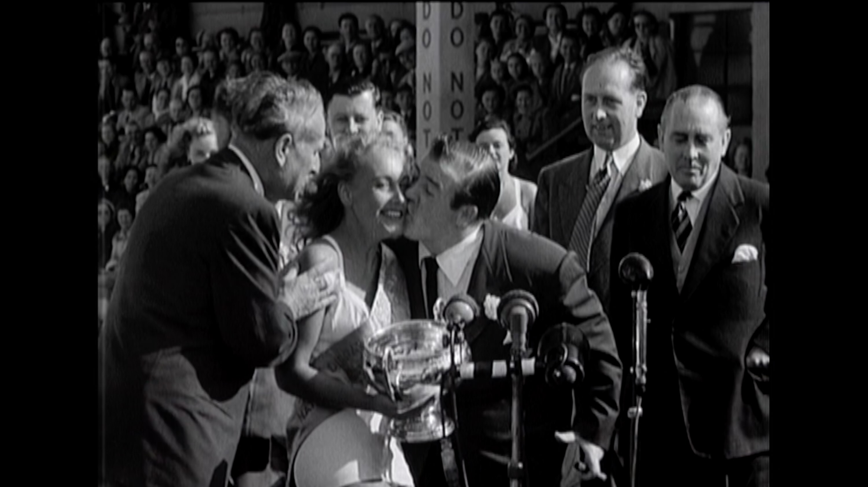 Still from music video black and white footage from 1950s beauty pageant awarding winner with trophy and kisses