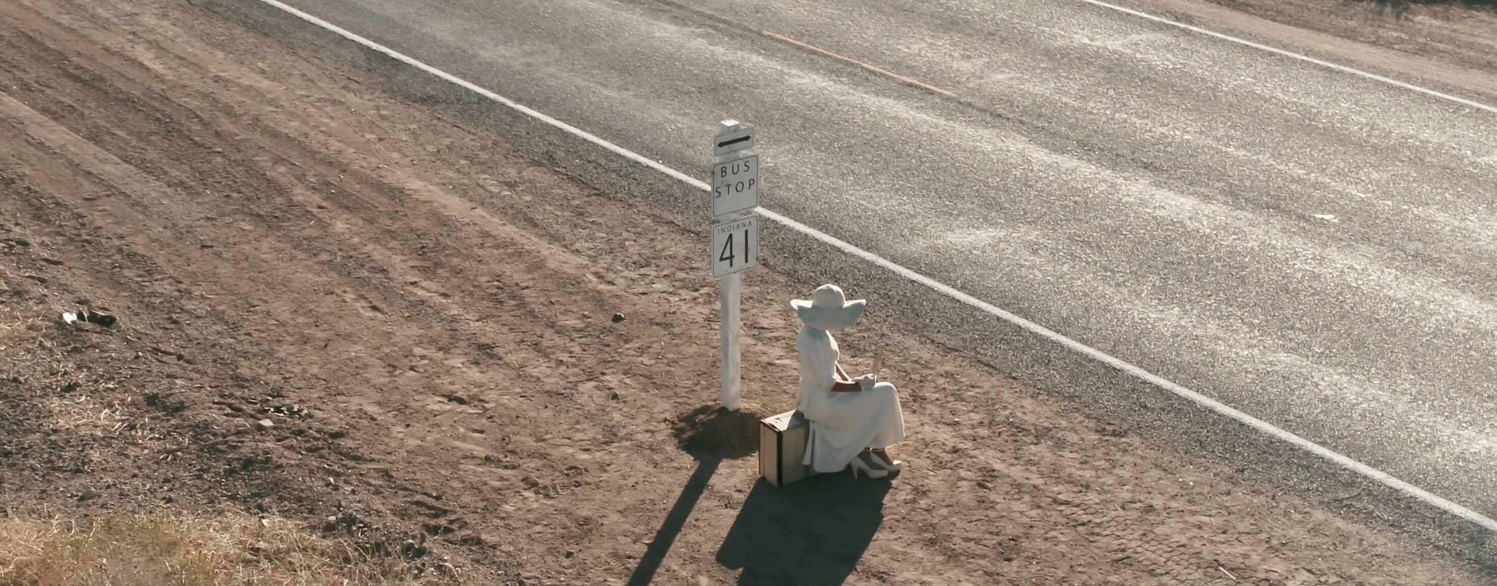 High angle still image from music video woman wearing large sunhat sitting on suitcase by bus stop at side of desert road