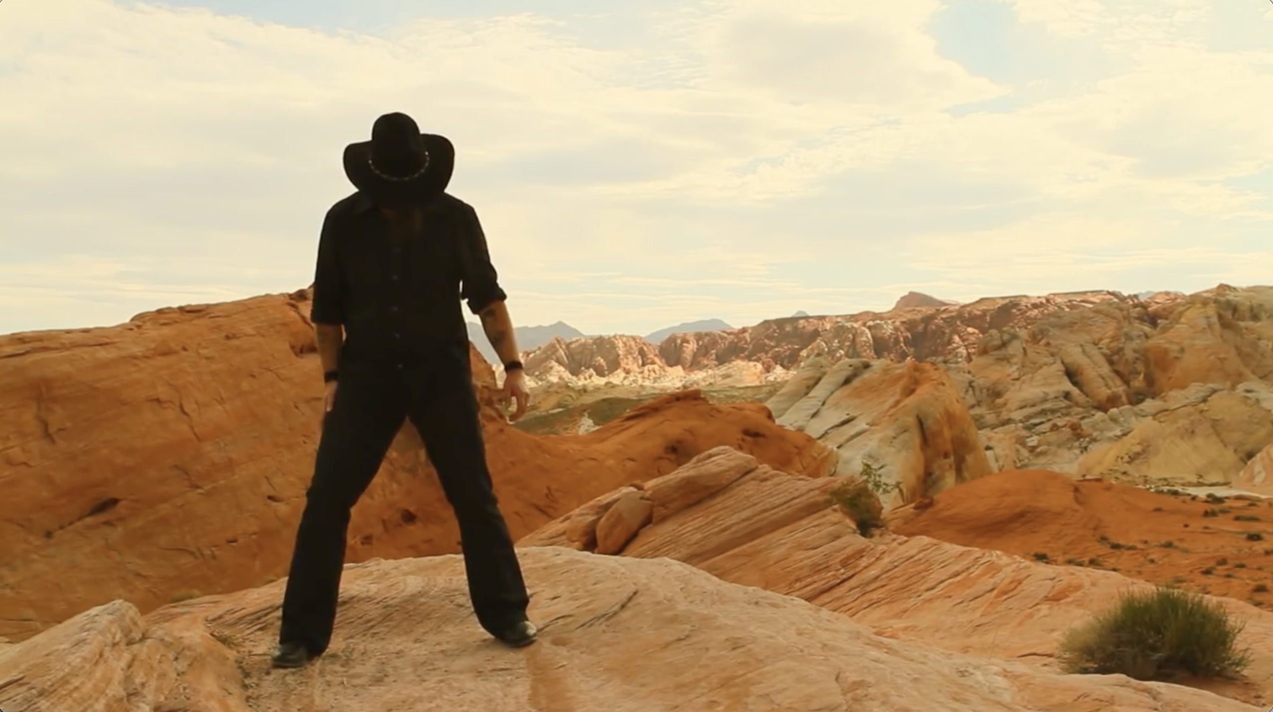 Still image from music video man dressed in black with black cowboy hat shielding his face in desert setting