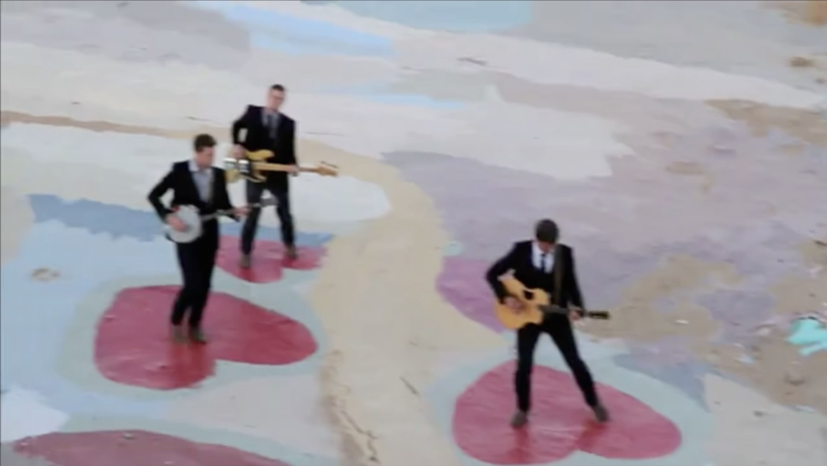 Music video still image trio wearing black suits standing in pink hearts painted on ground while playing instruments