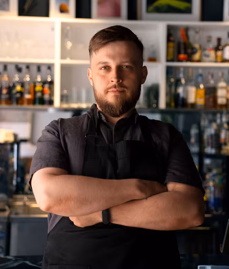 Bearded bartender in a black apron standing with arms crossed in front of a stocked bar
