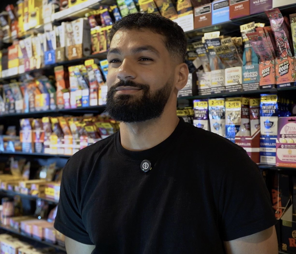 Man in a black t-shirt in front of tobacco product shelves at a smoke shop