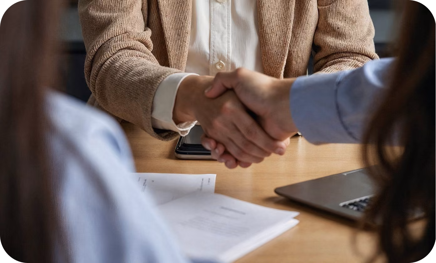 Two people shaking hands across a desk with resume and laptop during an interview