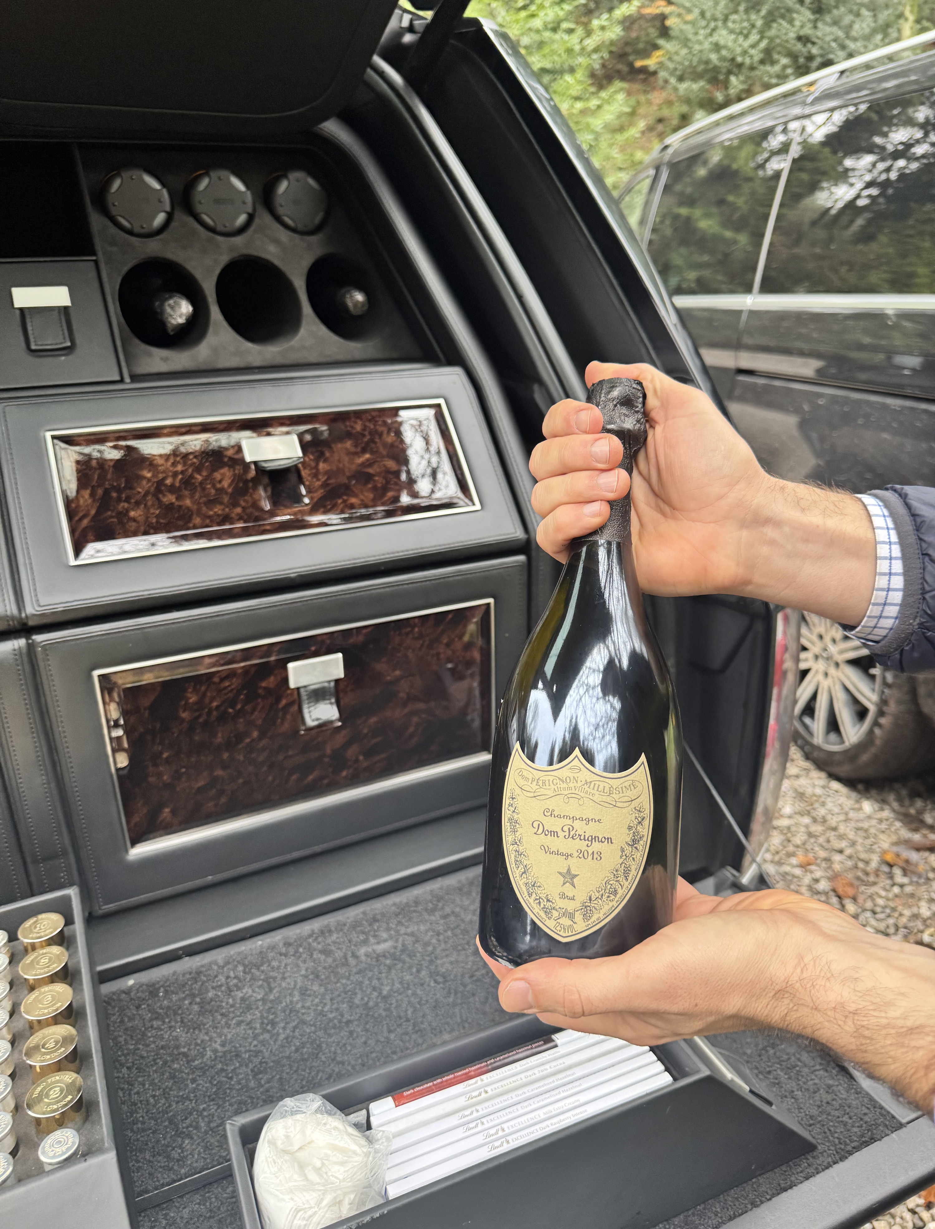A bottle of champagne held at the open bar cabinet of a Land Rover in the English countryside.