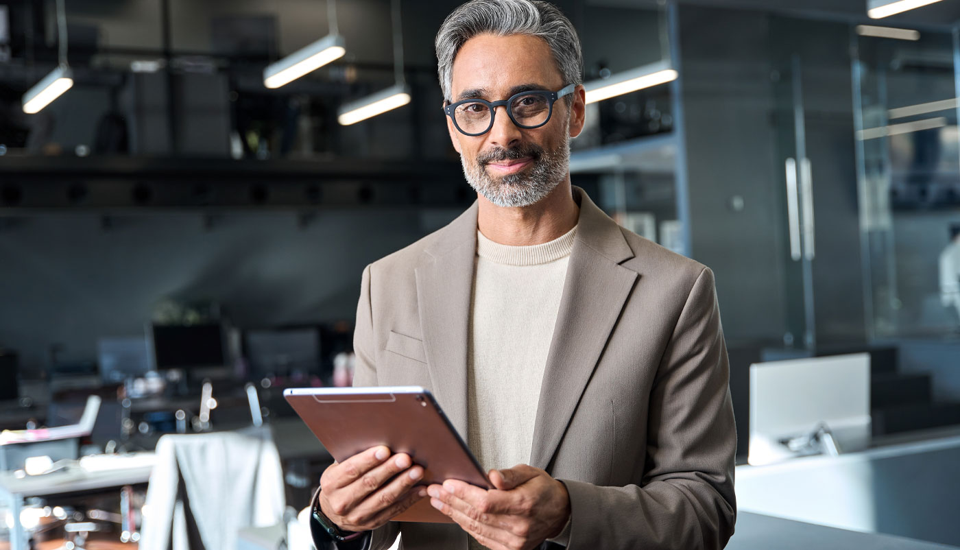 Smiling middle-aged man with gray hair and glasses holding a tablet in a modern office.