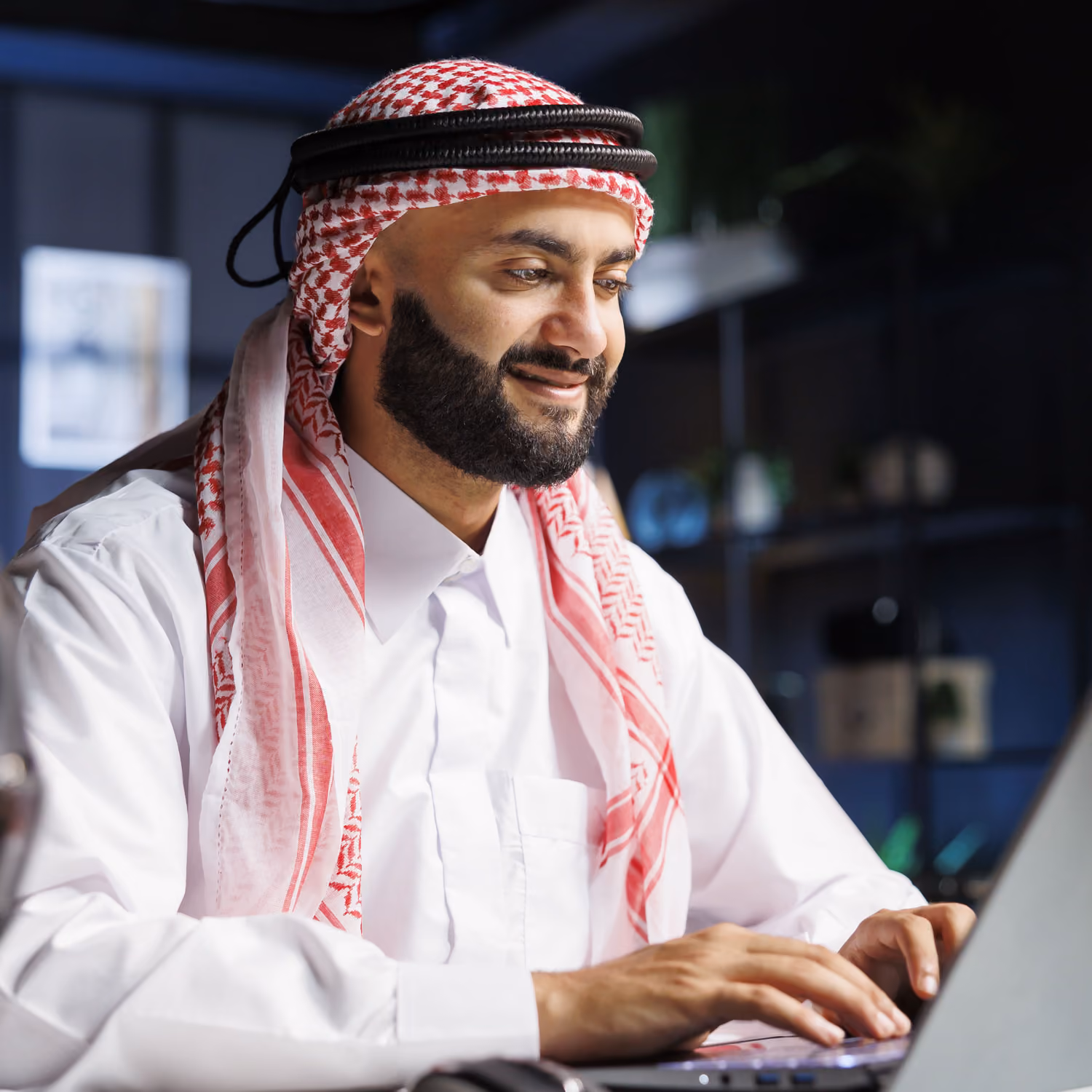 Man in traditional Middle Eastern attire smiling while typing on a laptop in a dimly lit room.