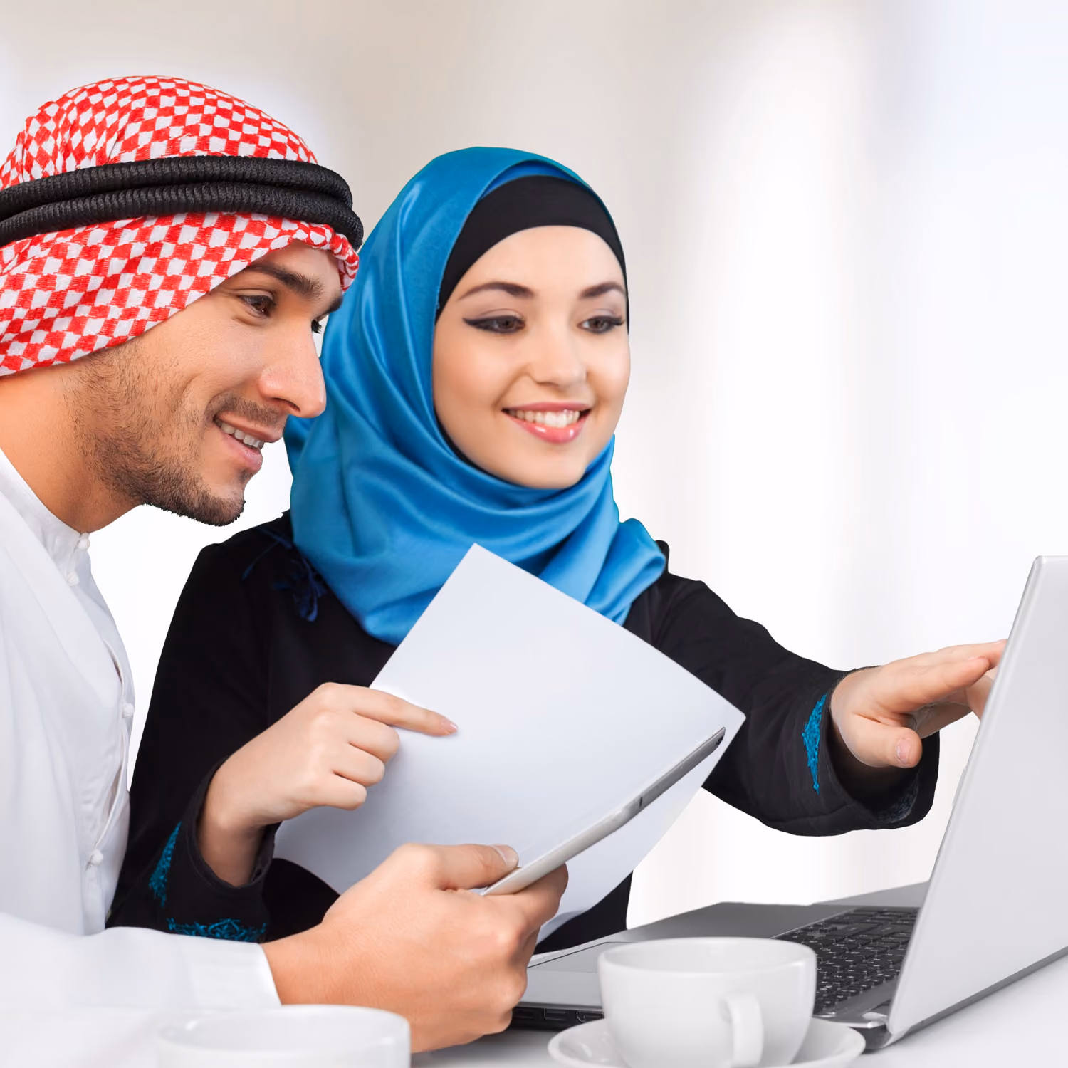 Two Arab people working together using a laptop and holding paper and tablet at a table with coffee cups.