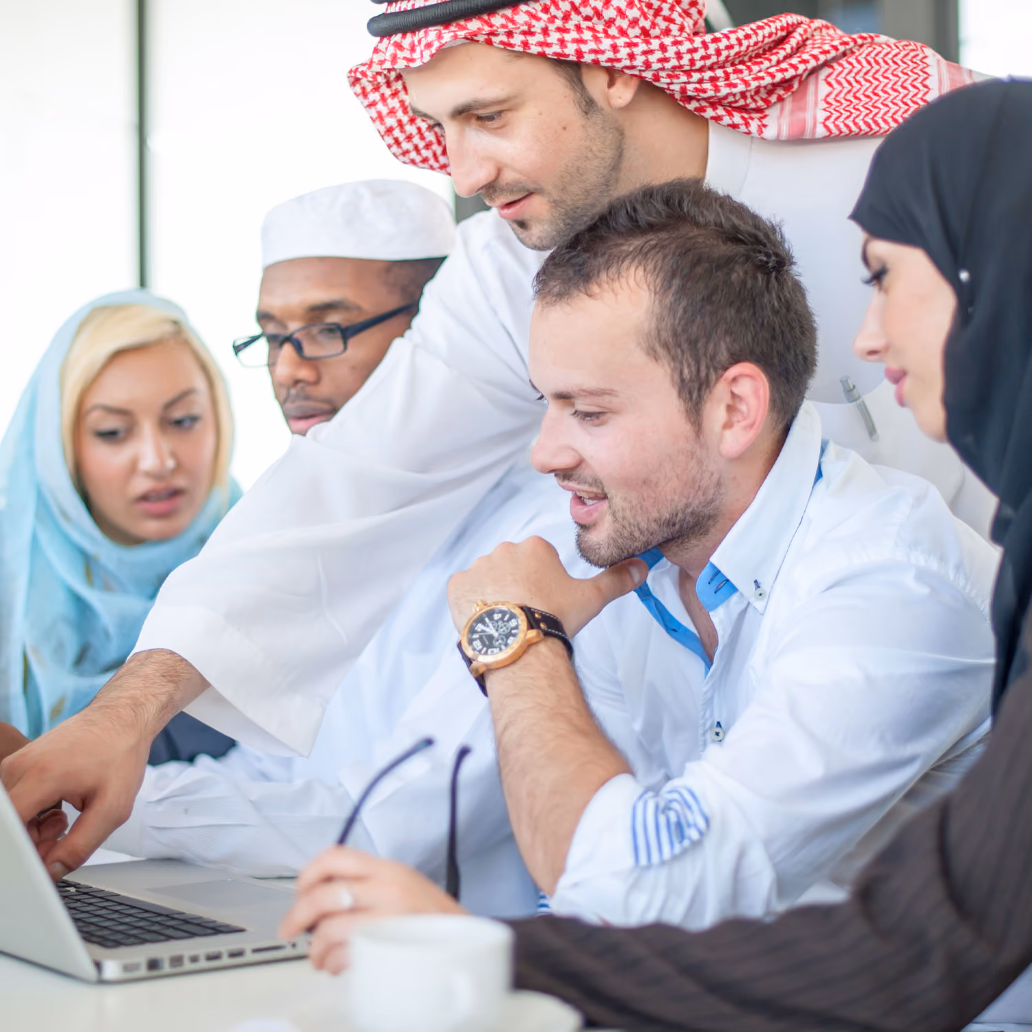 A diverse group of five people gathered around a laptop, collaborating and pointing at the screen.