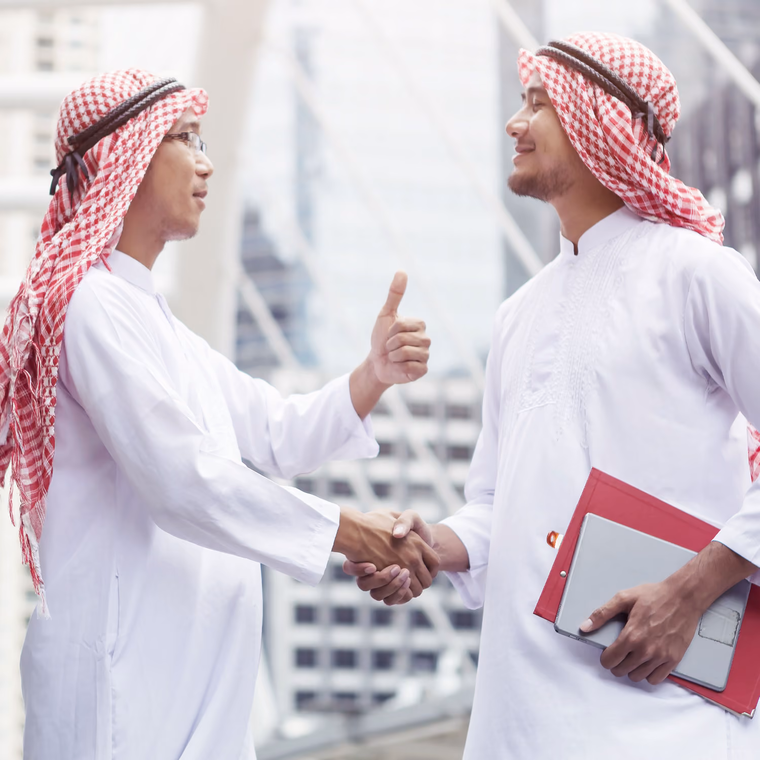 Two men in white thobes and red checkered headscarves shaking hands outside a modern building.