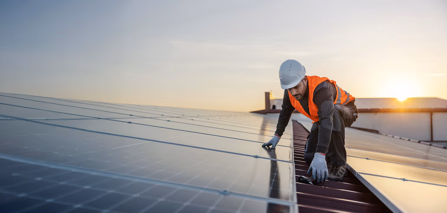 Worker in a safety helmet and high-visibility vest installing solar panels on a rooftop at sunset.