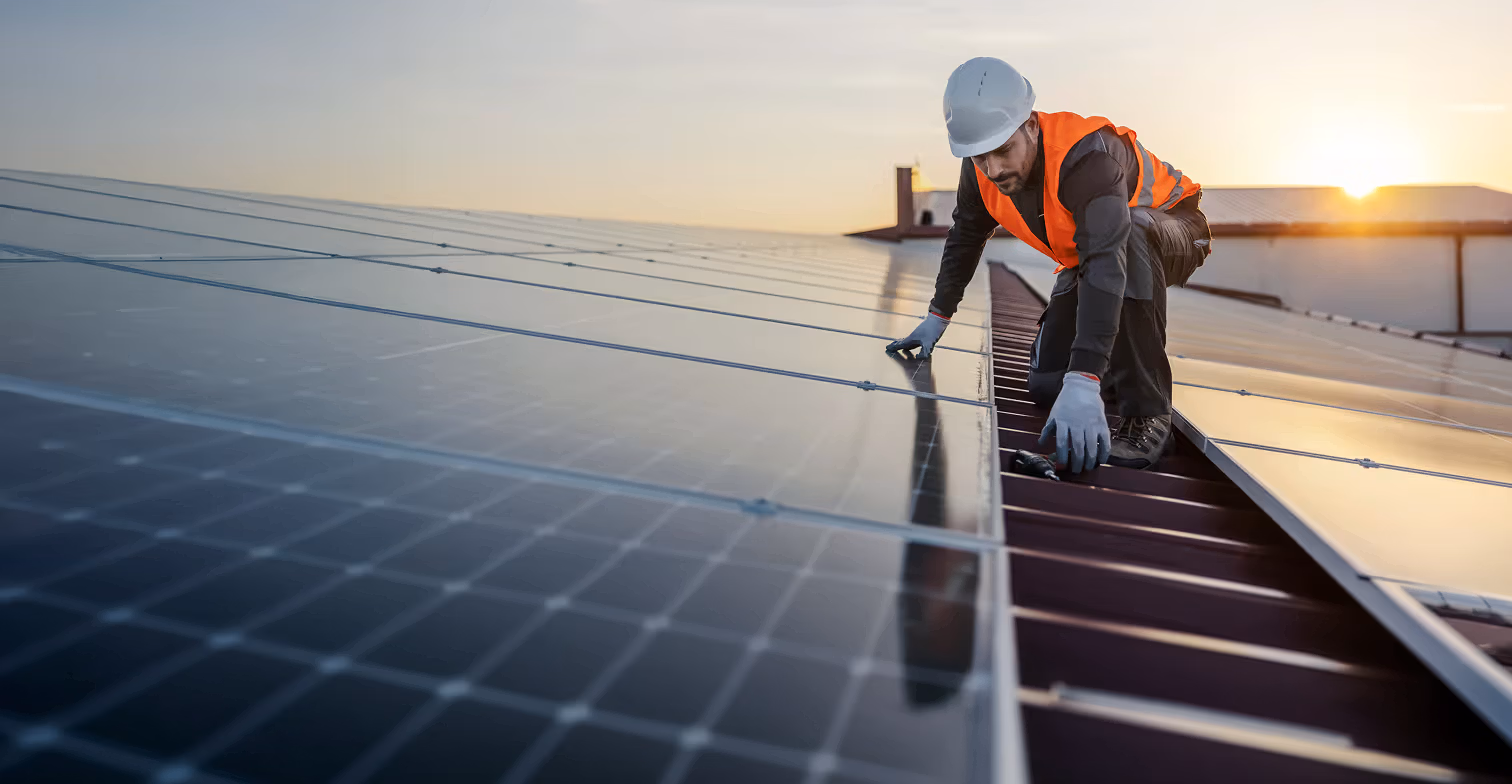 Worker in a safety helmet and high-visibility vest installing solar panels on a rooftop at sunset.