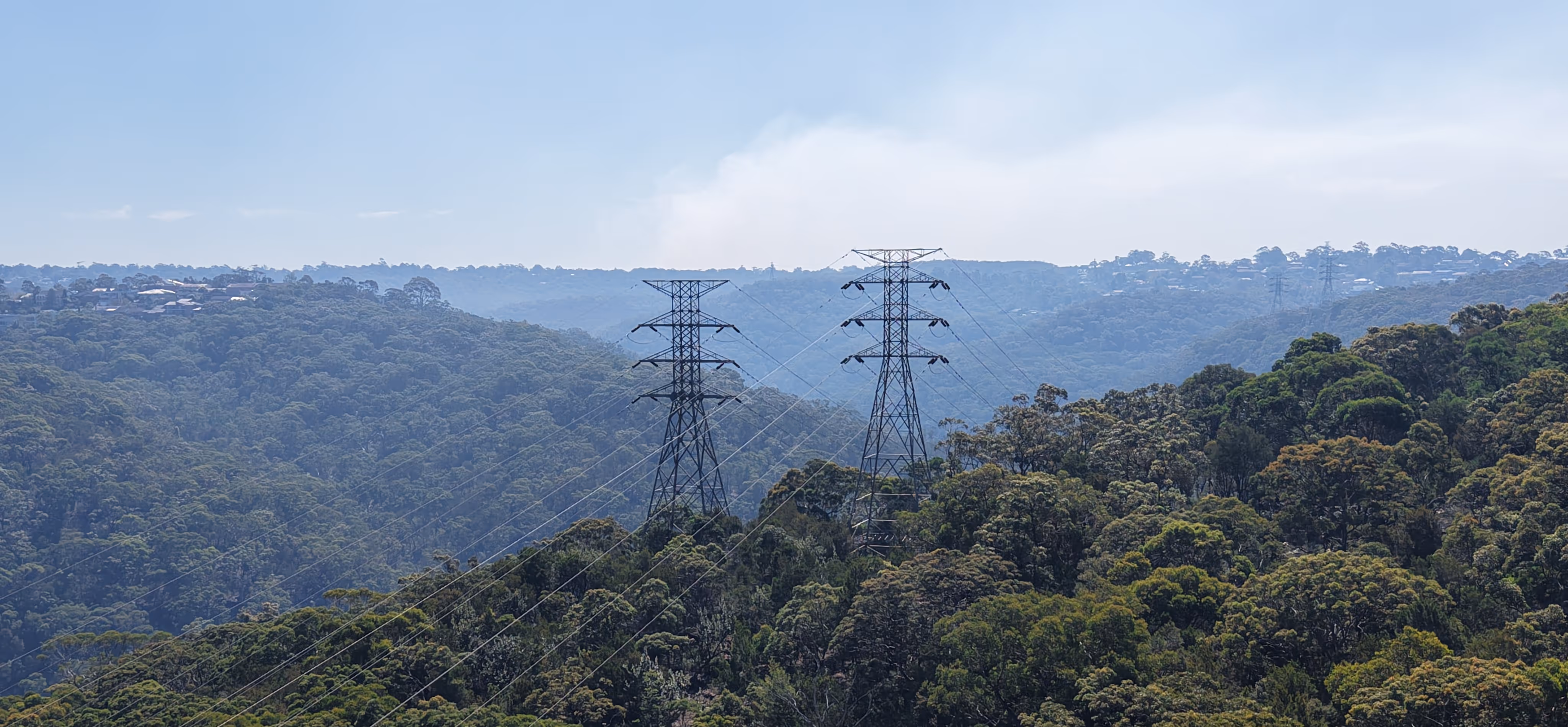 Two steel transmission towers with power lines stretching over a densely forested valley under a clear sky.