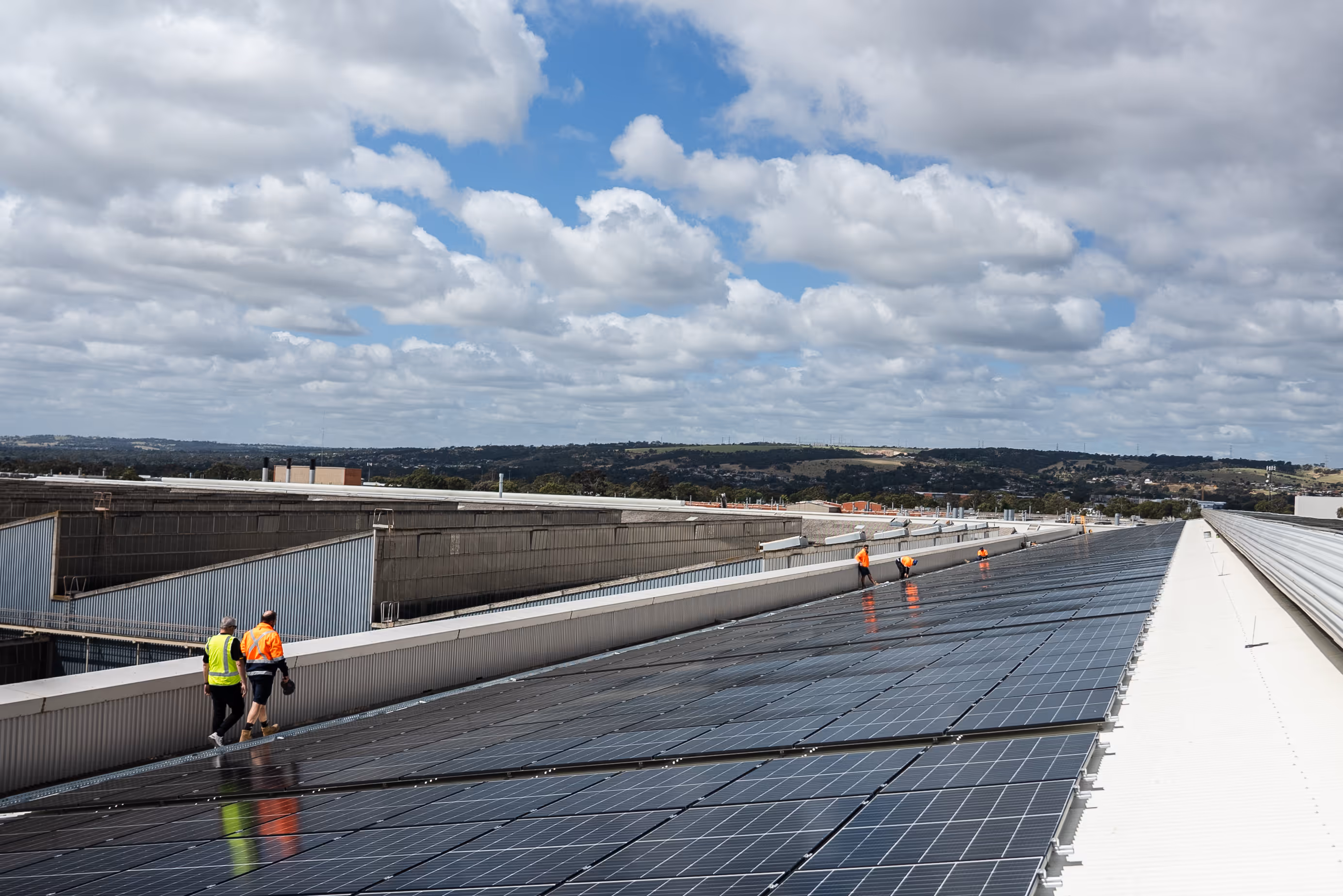Workers walking and inspecting large solar panel array on an industrial building roof under cloudy sky.