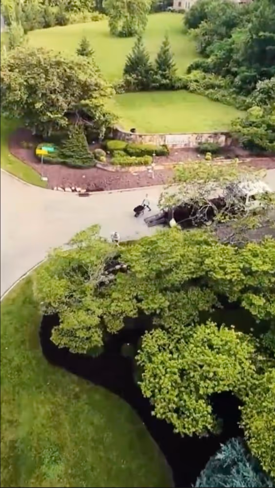 Aerial view of a suburban green area with trees, bushes, and a curved road where two people are walking near a parked vehicle.