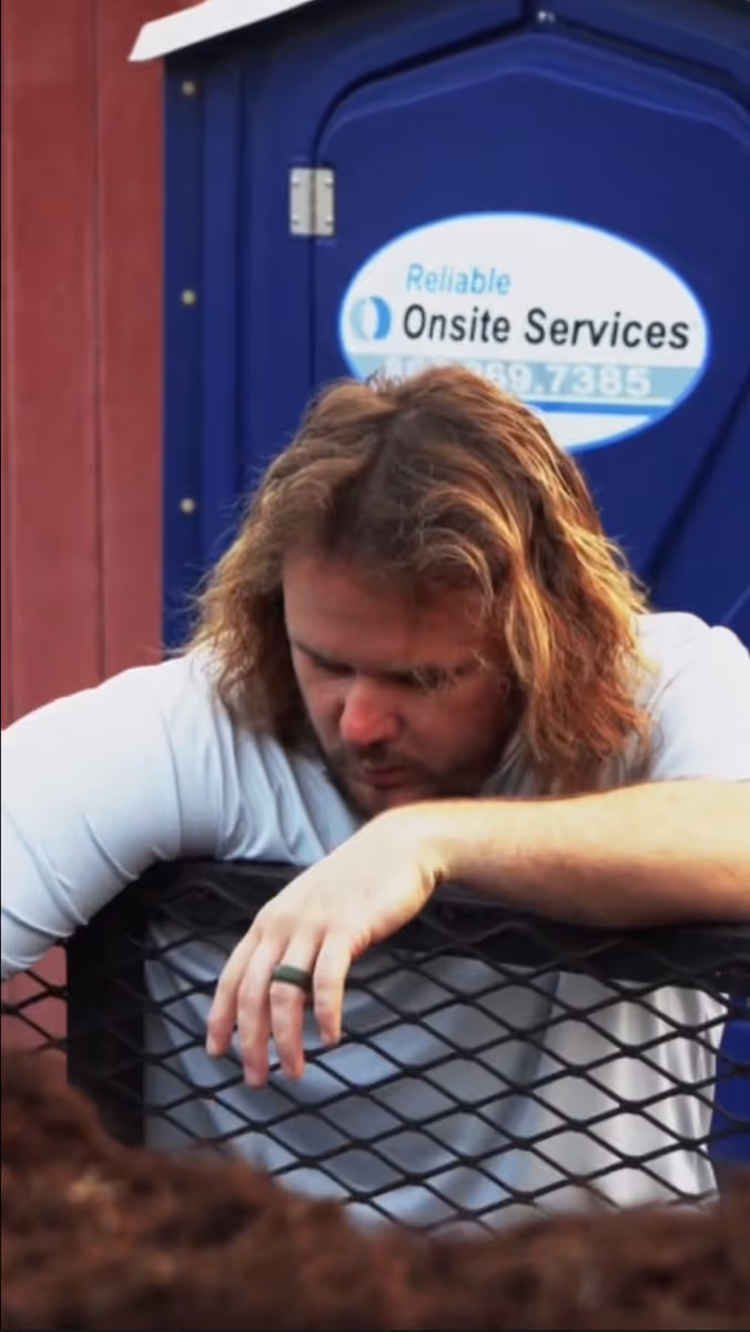 Man with long hair wearing a white shirt leaning on a black metal fence with a blue portable restroom behind him.