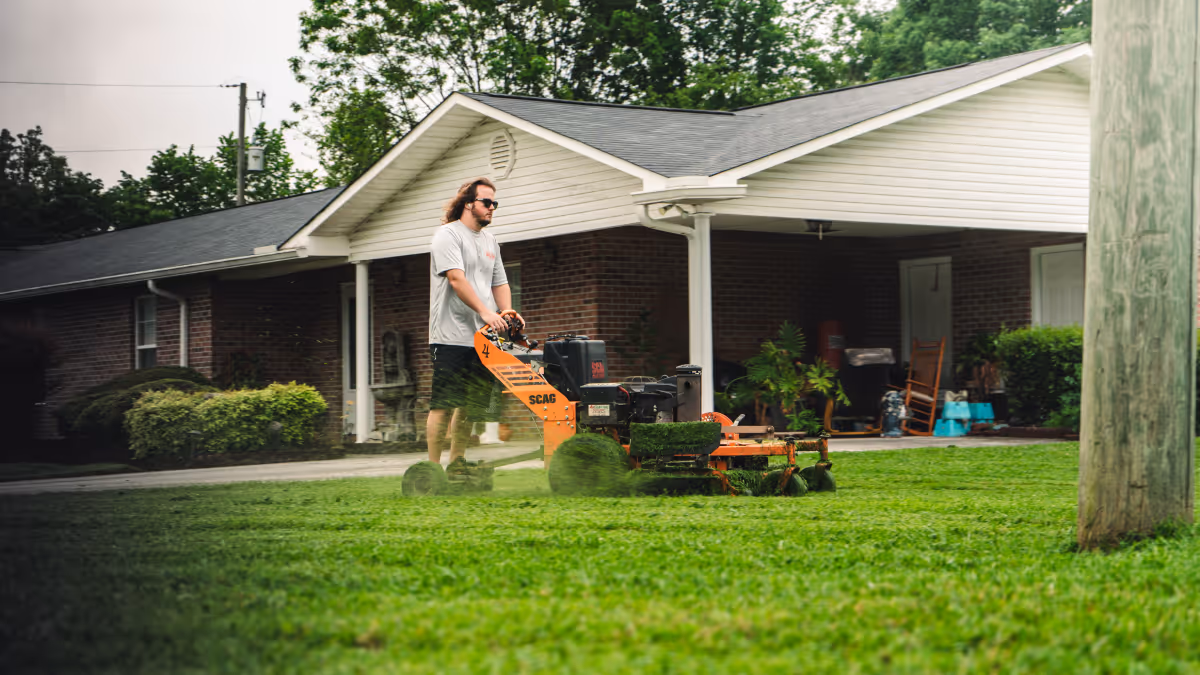 Man wearing sunglasses and shorts mowing a green lawn with an orange SCAG lawn mower in front of a brick house.
