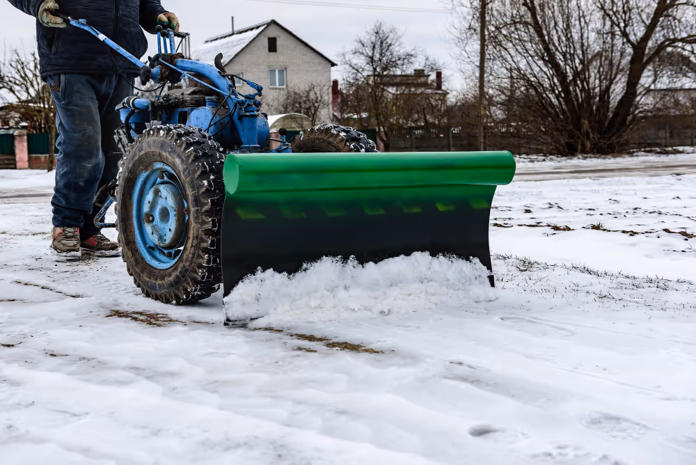 Person operating a blue snow plow machine clearing snow from a driveway with houses and leafless trees in the background.