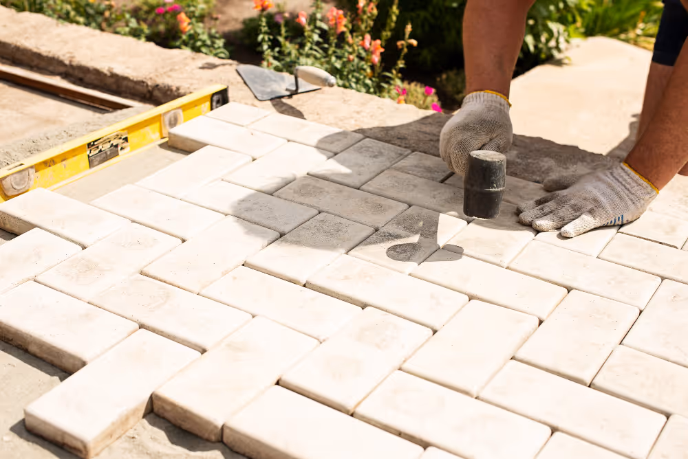 Worker wearing gloves installs rectangular white paving stones on a ground surface using a rubber mallet, with a level tool and flowers in the background.