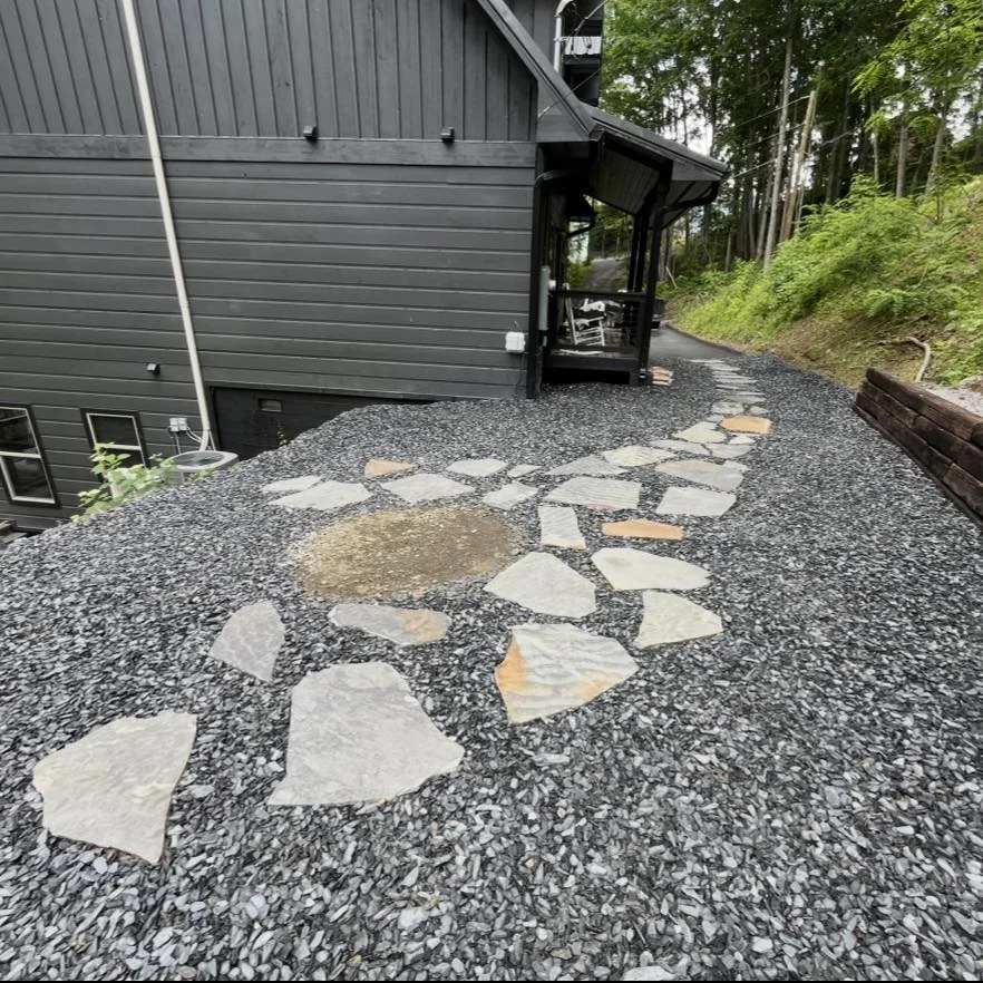 Gravel pathway with scattered large flat stones leading to a black house porch surrounded by green trees.