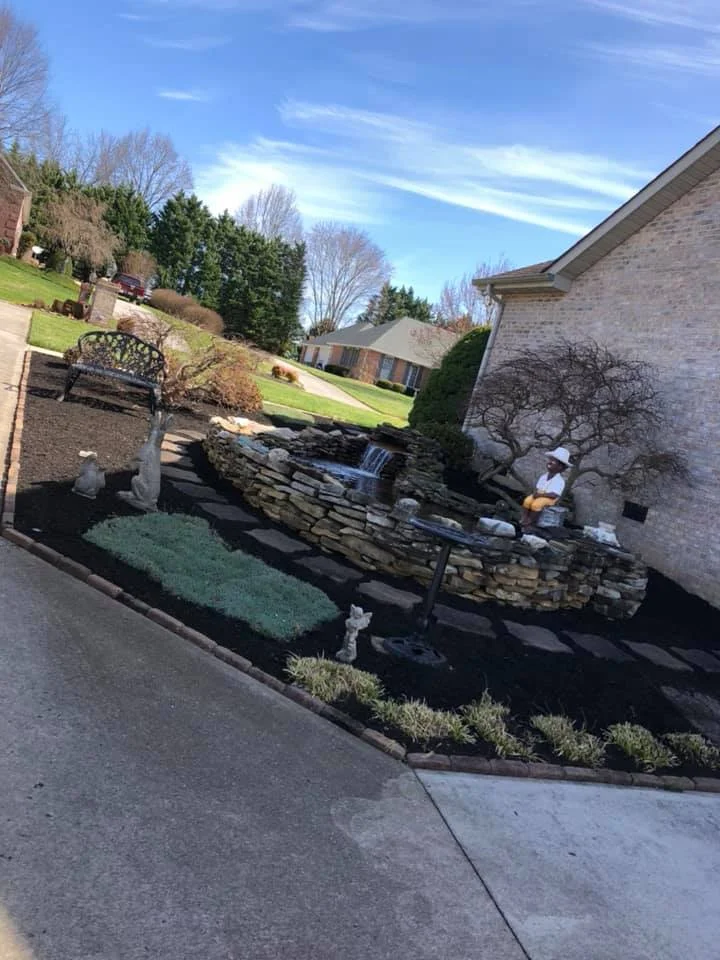 Residential front yard with a stone-lined pond and waterfall, decorative statues, black mulch, and a bench under a clear blue sky.