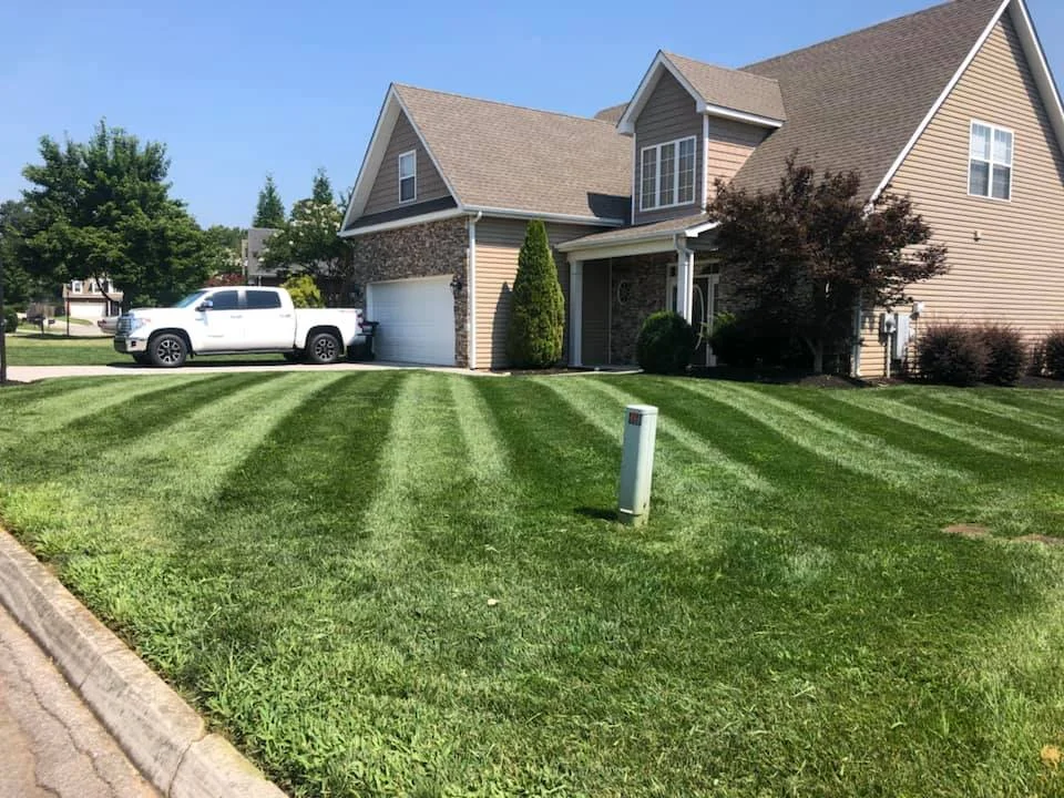 Well-maintained lawn with striped mowing pattern in front of a beige two-story house with a white pickup truck parked in the driveway.