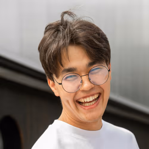 Smiling young man with round glasses and tousled hair wearing a white shirt, against a blurred outdoor background.