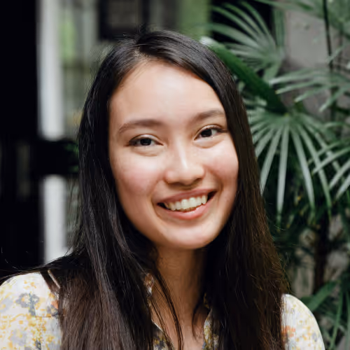 Young woman with long dark hair smiling in front of green plants indoors.