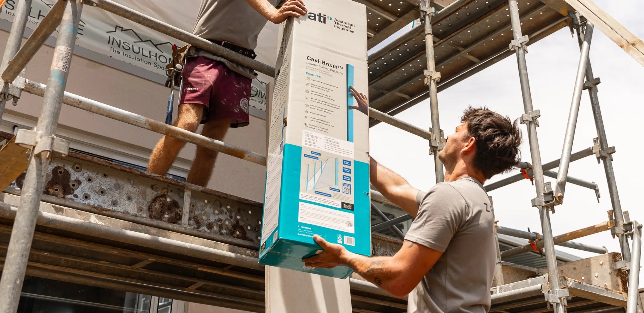 Two construction workers on scaffolding handling a large box of Australian Thermal Industries' Cavi-Break insulation product.