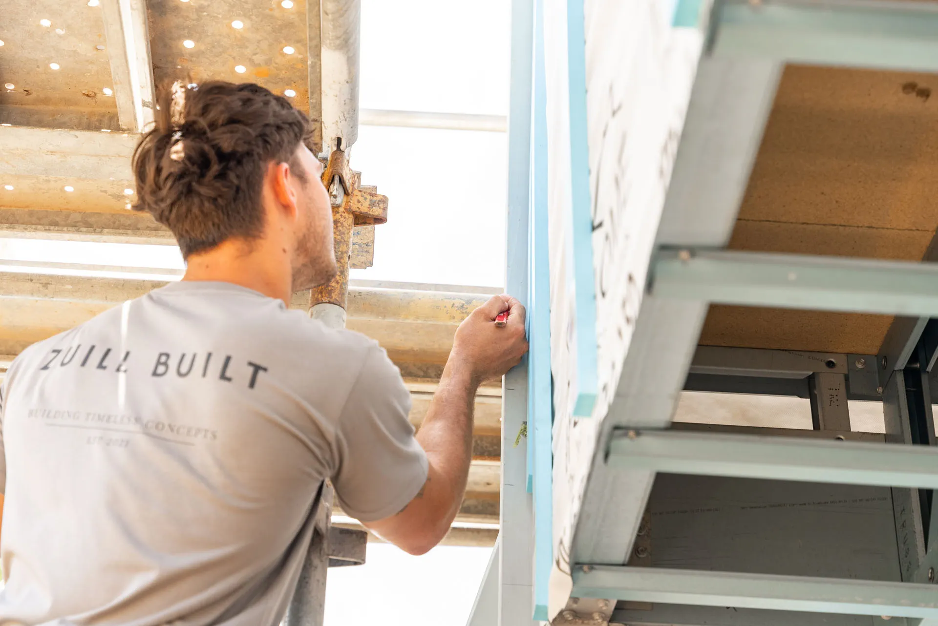 Construction worker in a grey shirt marking a light blue wall near scaffolding on a building site.