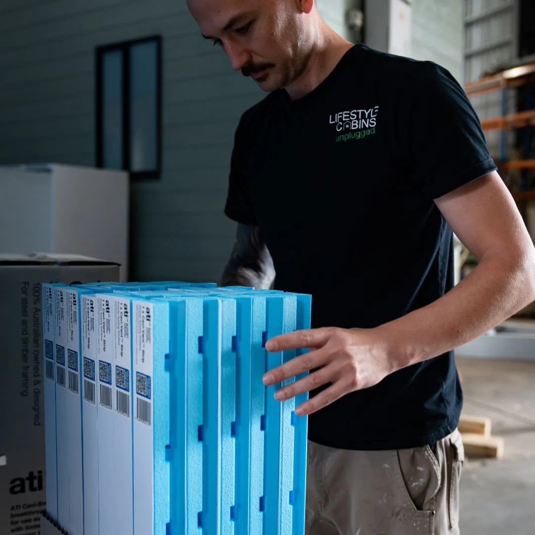 Man in black Lifestyle Cabins shirt handling blue and white insulated construction panels indoors.