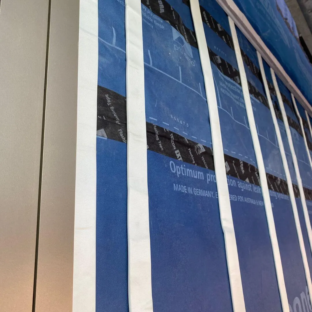 Wall under construction with blue weatherproof barrier and white tape strips applied vertically.