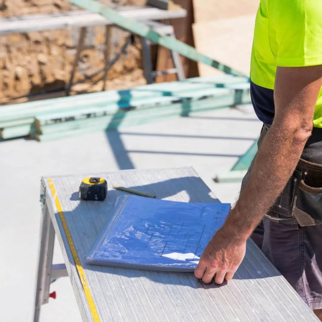 Construction worker wearing a yellow shirt reviewing blueprints on a workbench at a building site.