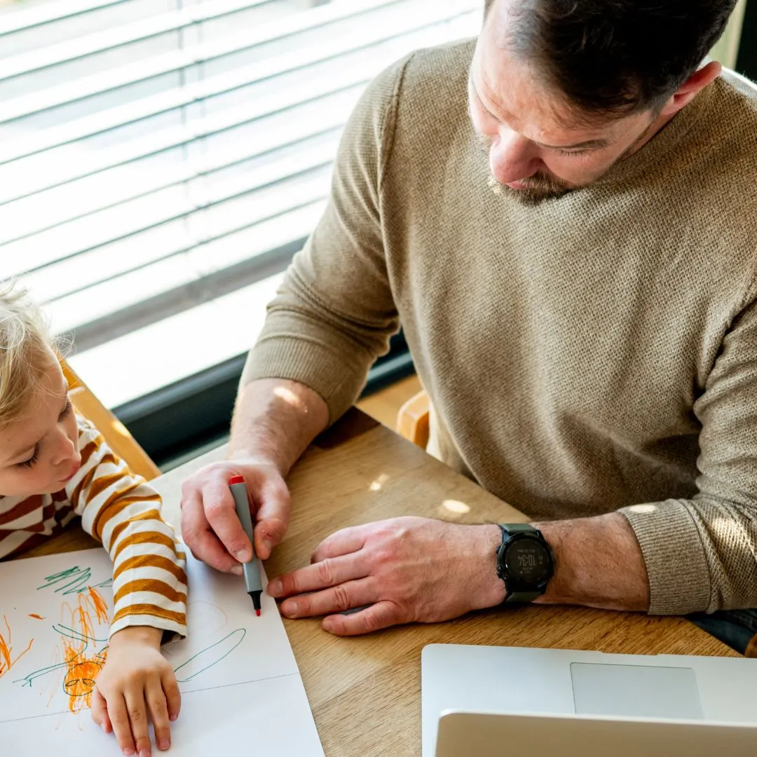 Man and child drawing with markers on paper at a wooden table near a laptop.
