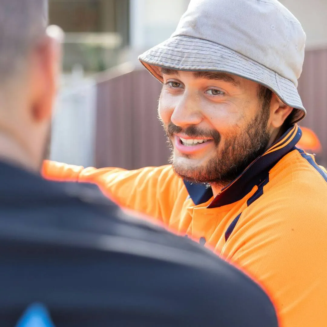 Man in orange work shirt and bucket hat smiling and talking to another person.