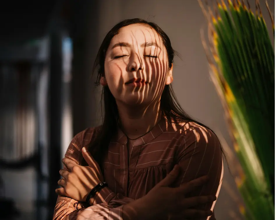Woman with closed eyes embracing herself as leaf shadows fall across her face and body.