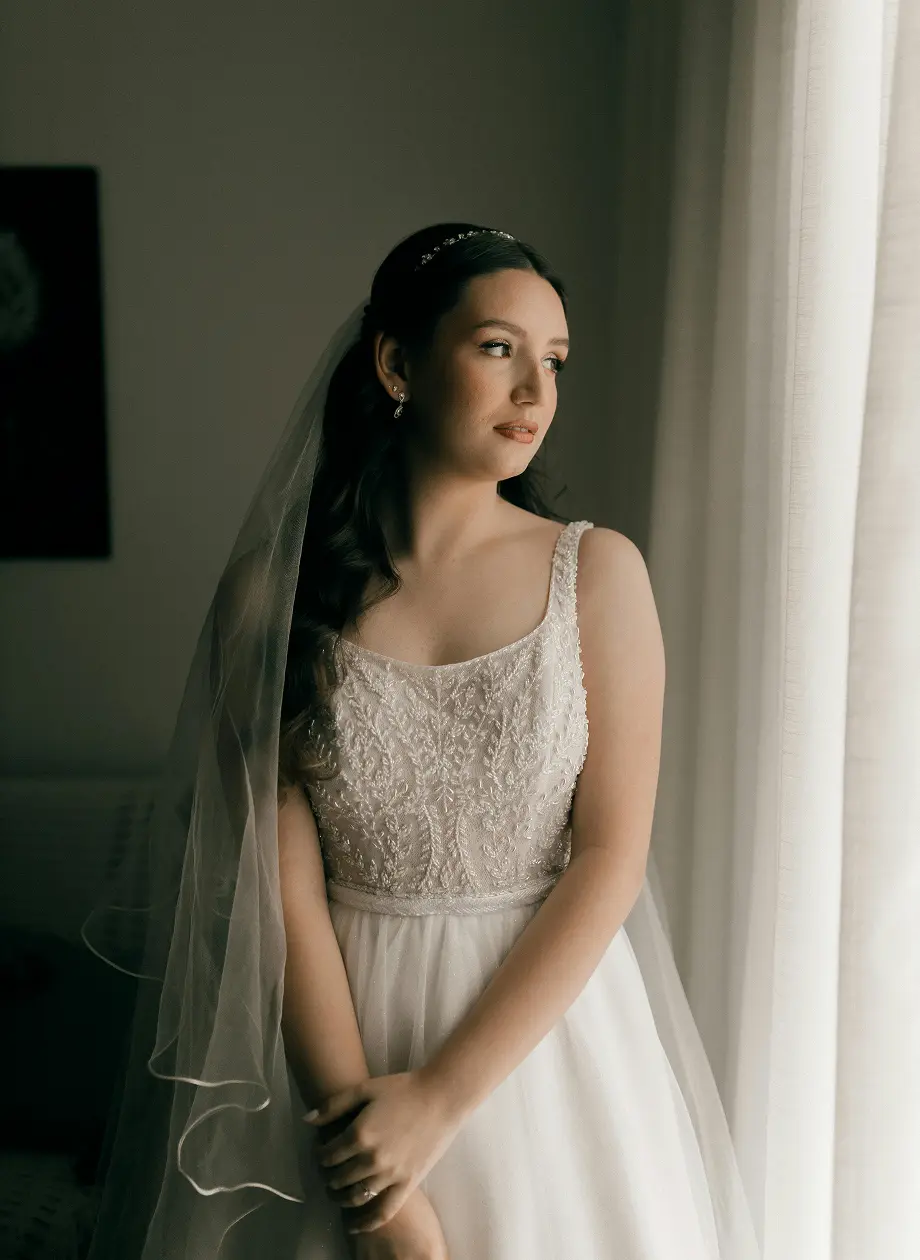Bride in an embroidered white gown and veil looking thoughtfully through a sheer curtained window.