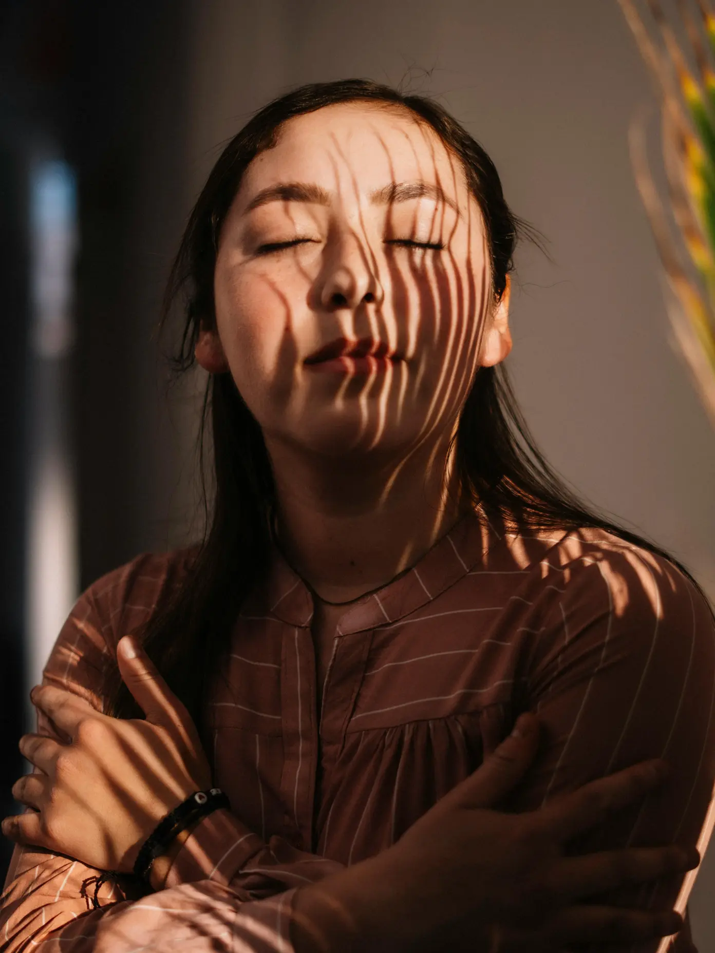 Woman with closed eyes and arms crossed as striped shadows fall across her face and clothes.