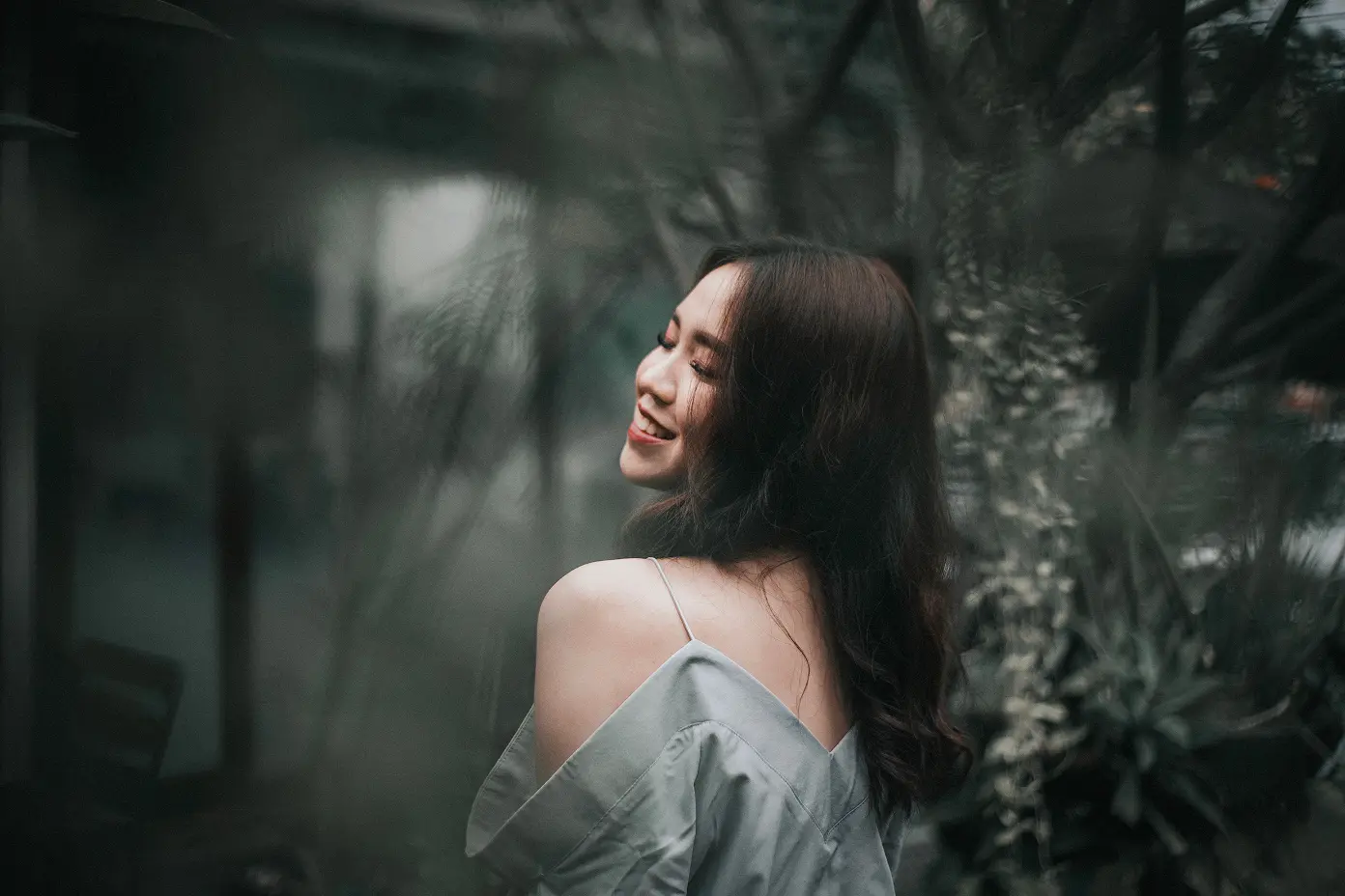 Smiling woman with long dark hair and off-shoulder top in an outdoor garden setting.