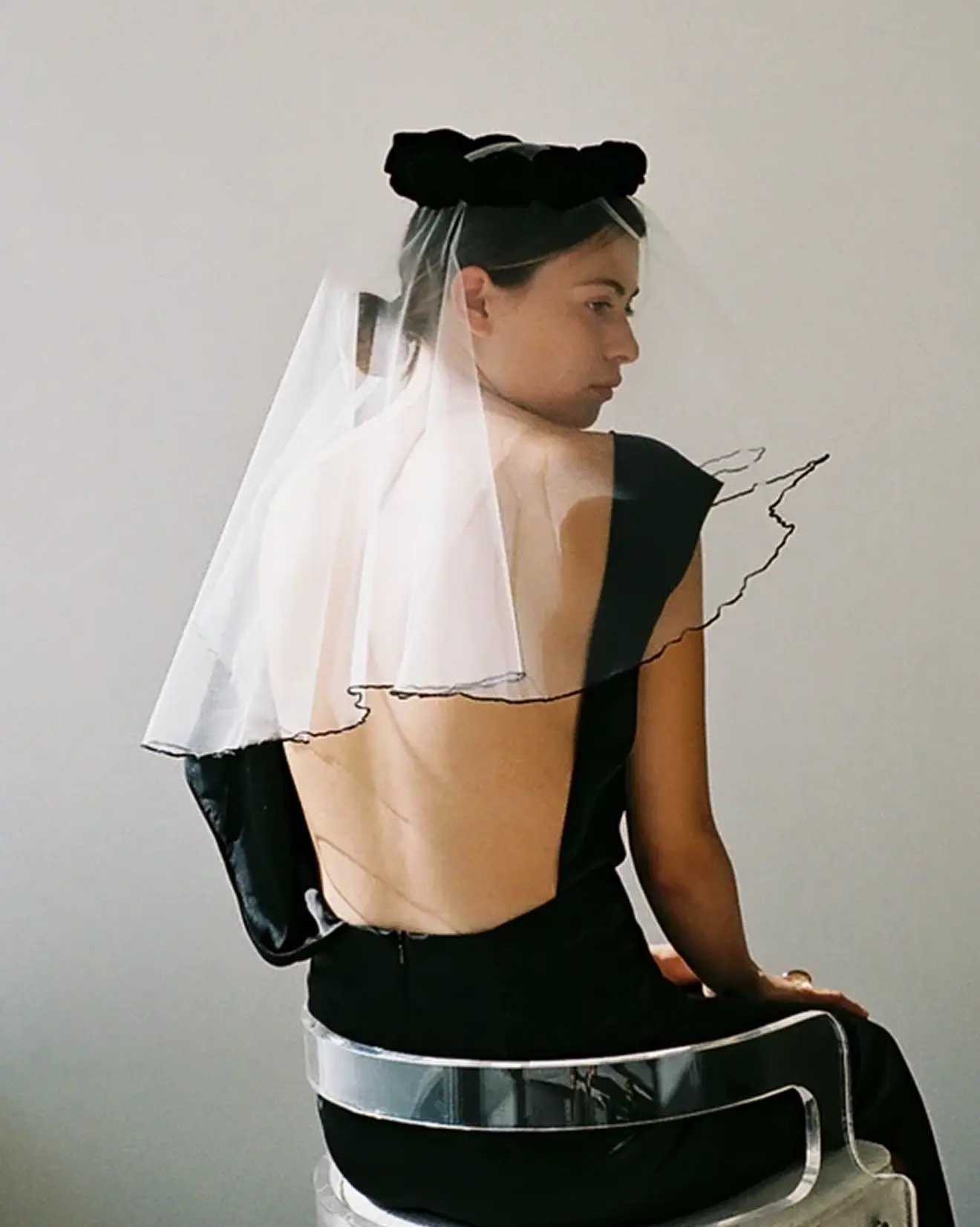 Woman in a black backless dress wearing a veil and sitting on a clear acrylic chair.