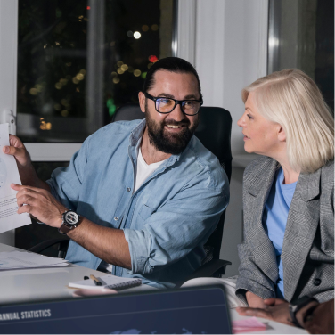 A man and woman in business attire discussing and pointing at a document in an office at night.