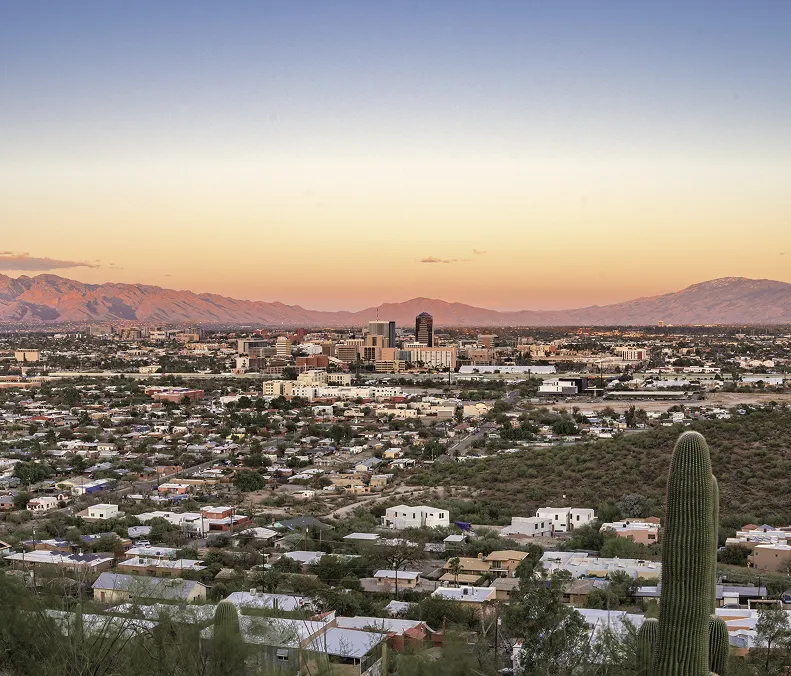 Cityscape at sunset with a desert foreground, houses, and mountains in the background under a clear sky.