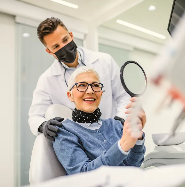 Smiling older woman holding a round mirror in a dentist's chair while a male dentist wearing a black mask and gloves stands behind her.