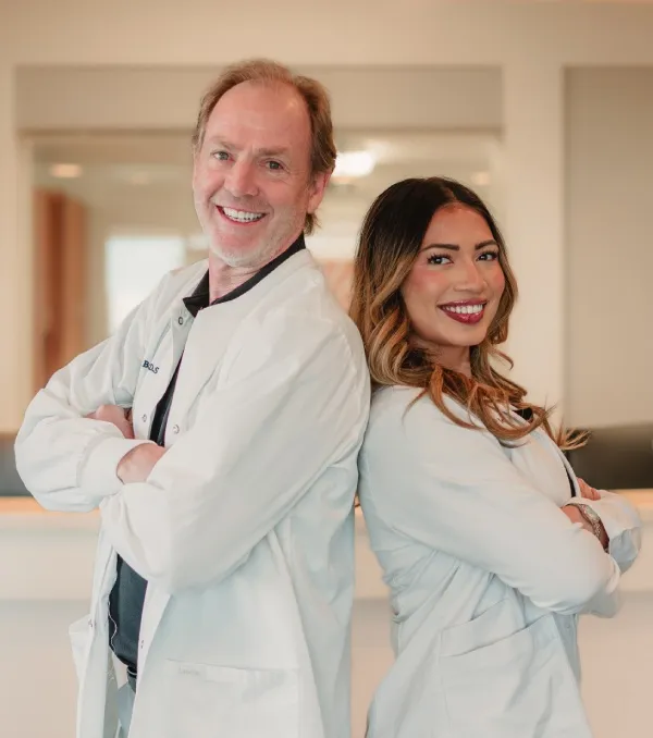 Male and female medical professionals standing back to back with arms crossed, smiling in a clinical setting.