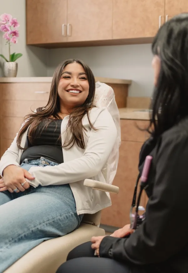 Smiling woman in casual clothes sitting in a dental chair talking to a healthcare professional.