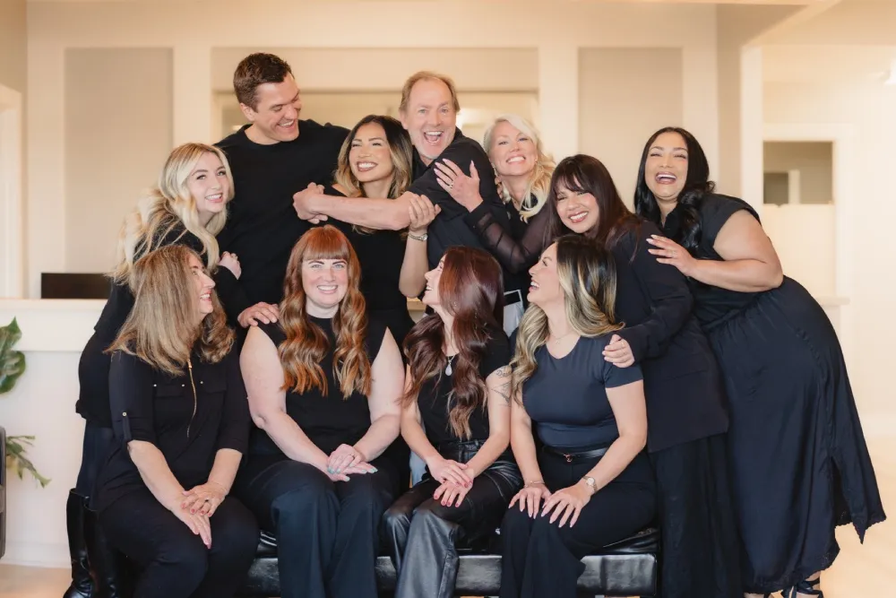 Group portrait of ten people smiling and embracing in an indoor setting with light-colored walls.