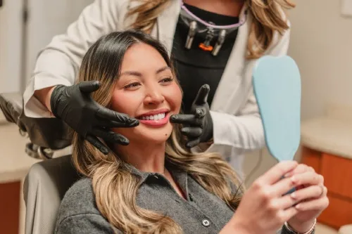 Smiling woman sitting in dental chair holding a mirror, while dentist wearing black gloves examines her face.
