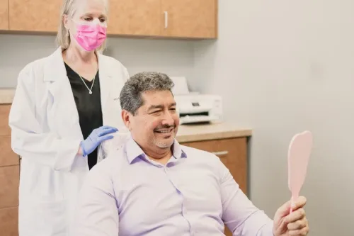 Smiling man sitting in dental office looking at a handheld mirror while a female dentist in a pink mask stands behind him.