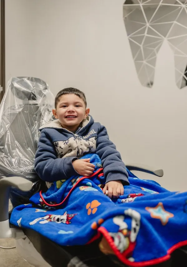 Smiling young boy sitting in a dental chair covered with a blue blanket featuring cartoon dog characters.