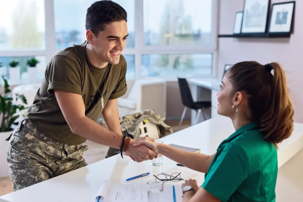 Smiling soldier shaking hands with a woman in a green shirt across a desk with paperwork and glasses.