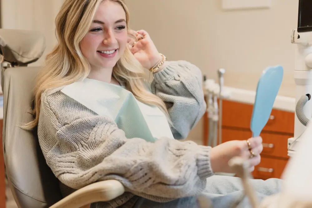 Smiling young woman with blonde hair sitting in a dental chair looking at a blue hand mirror while wearing a dental bib.